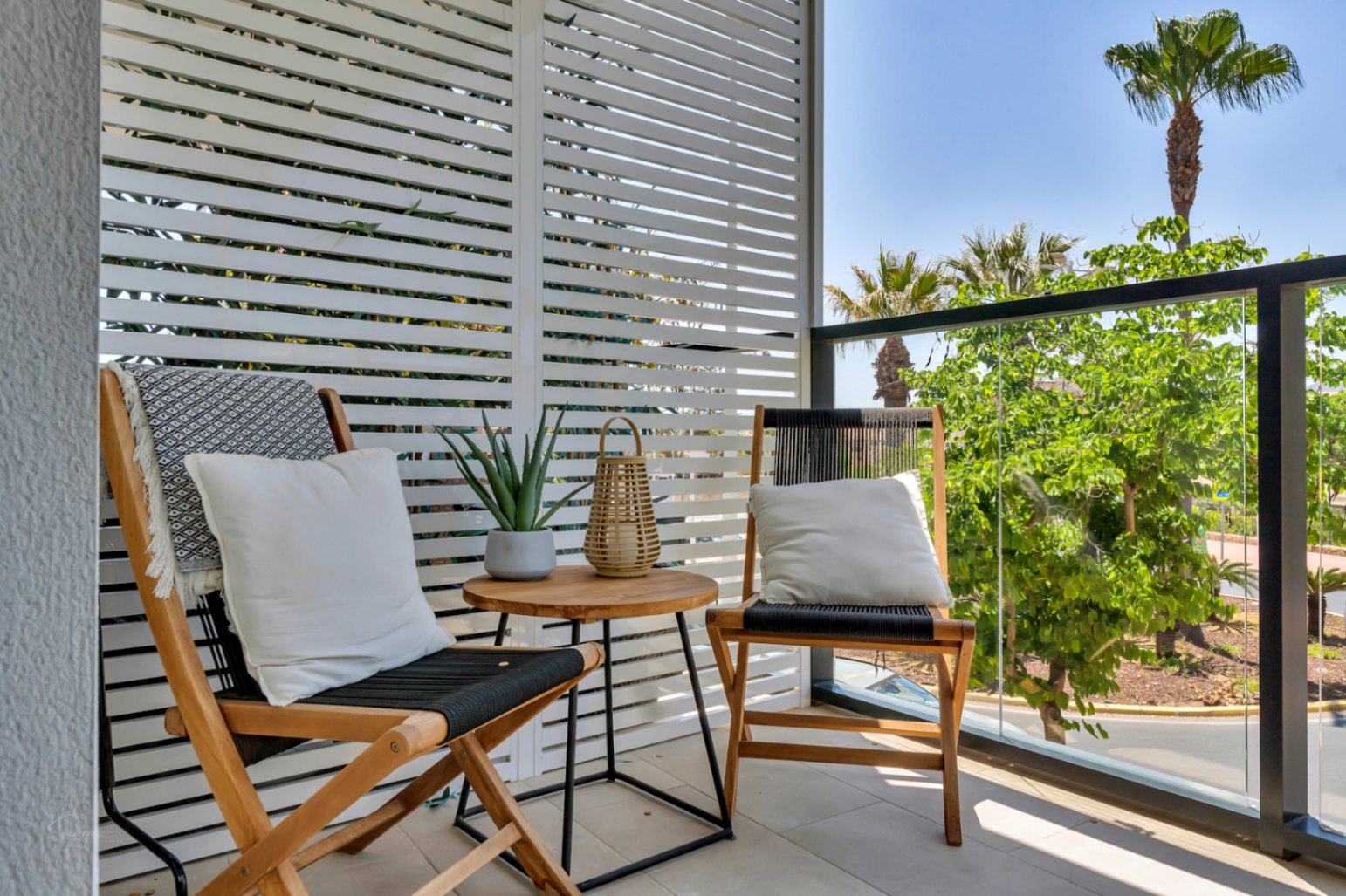 Terraza privada con cerramiento de lamas blancas, mobiliario de madera y vistas despejadas a zonas verdes con palmeras bajo el cielo mediterráneo.