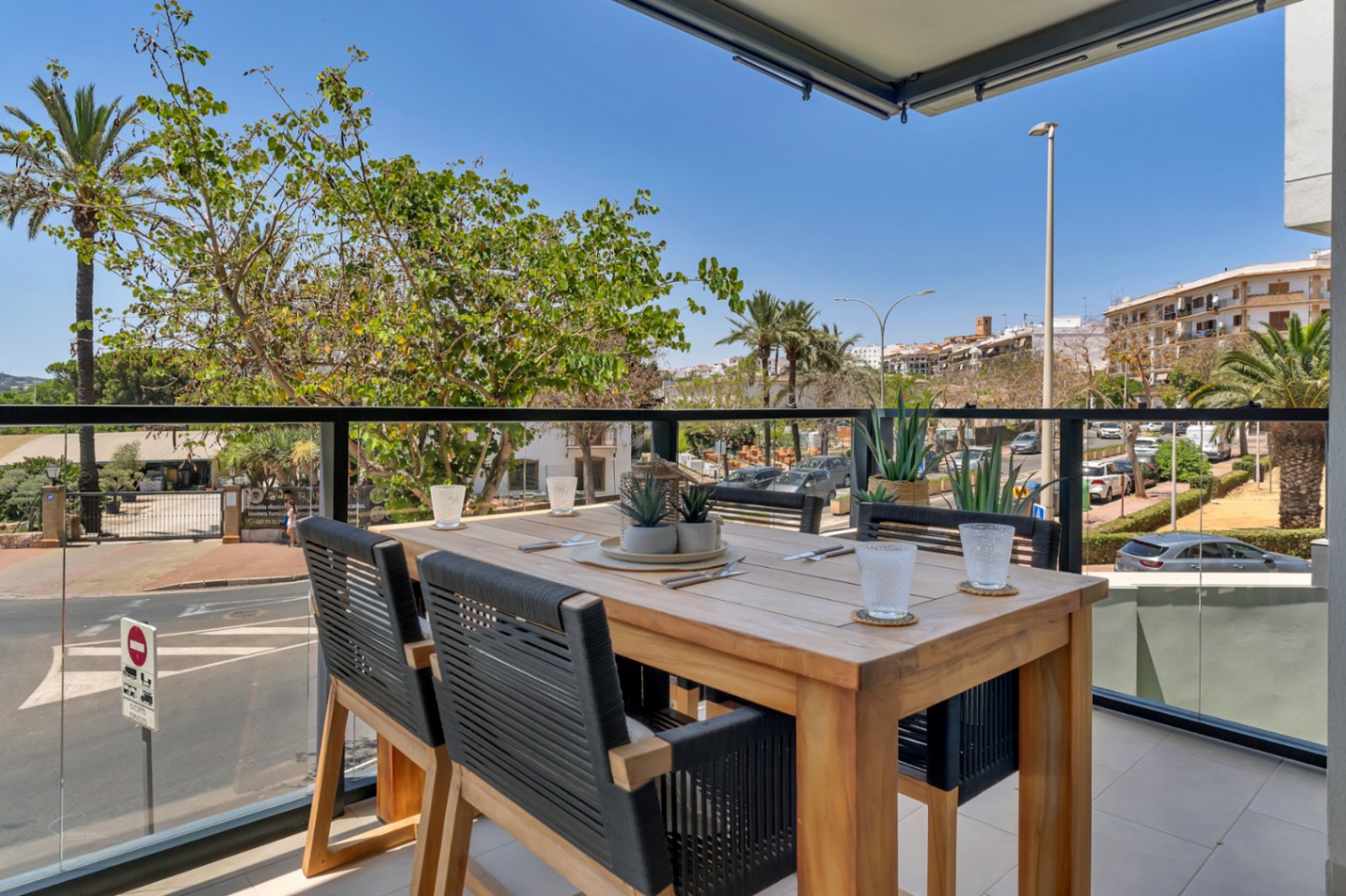 Terraza privada con mesa de comedor de madera, sillas de cuerda trenzada y barandilla de cristal con vistas despejadas al entorno urbano y palmeras.