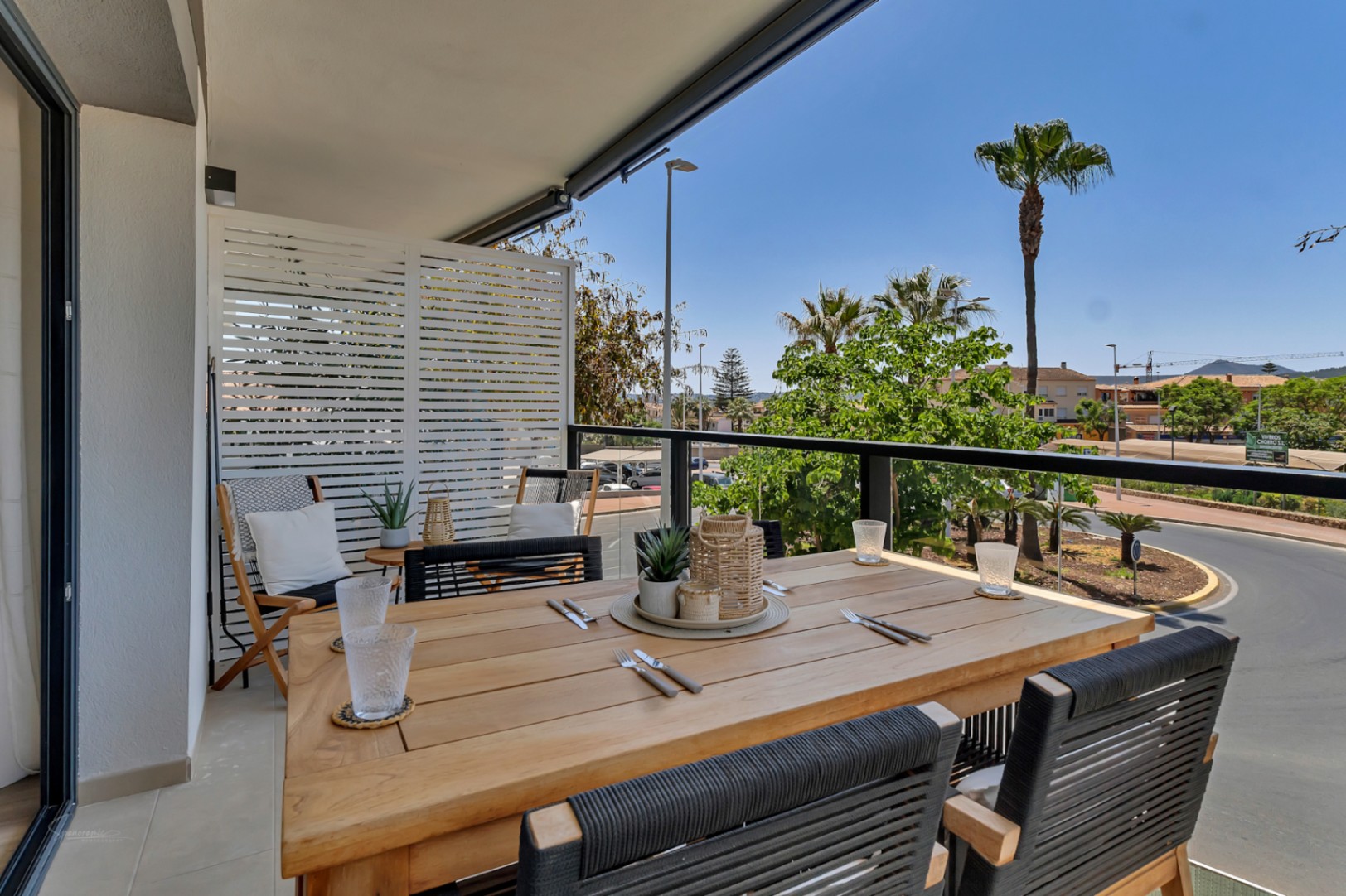 Terraza privada con mesa de comedor de madera, sillas de cuerda trenzada y barandilla de cristal con vistas despejadas y palmeras en Jávea.