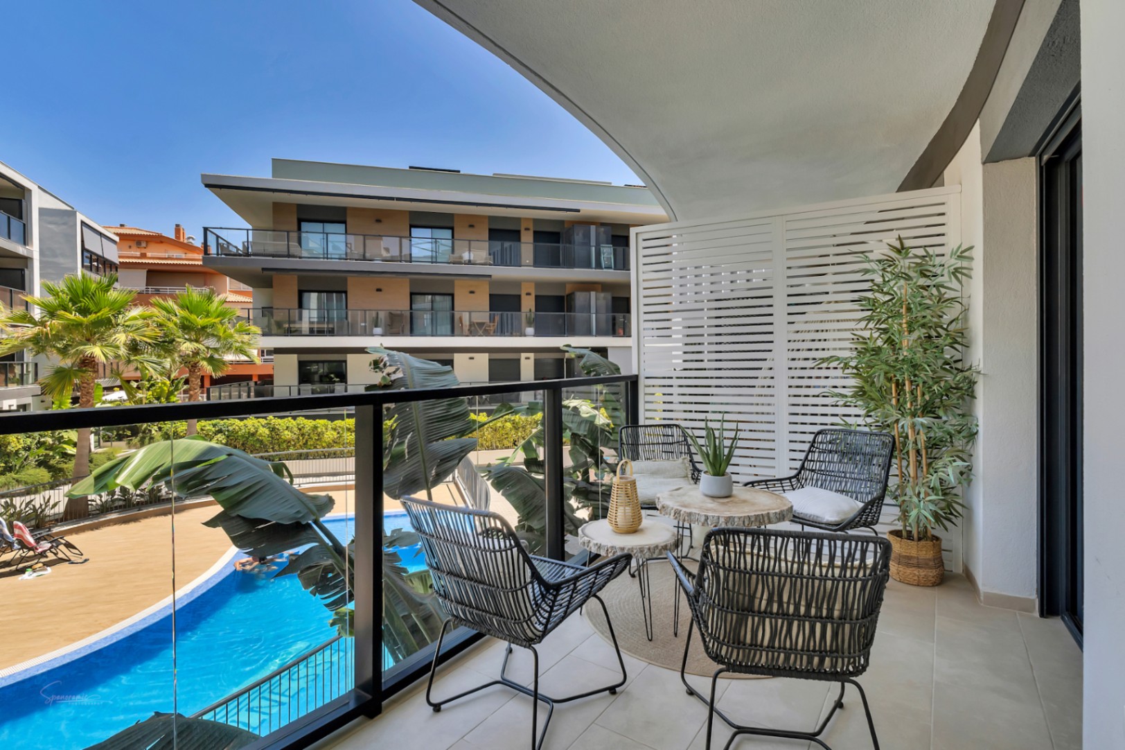 Terraza privada con mobiliario de diseño, barandilla de cristal y vistas directas a la piscina comunitaria y zonas ajardinadas bajo un cielo despejado.