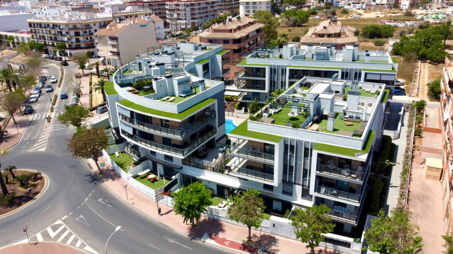 Vista aérea de un complejo residencial contemporáneo en Jávea, con fachadas curvas, amplias terrazas con césped artificial y una piscina comunitaria central.