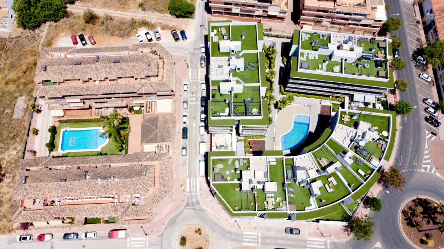 Vista aérea del complejo residencial contemporáneo en Jávea, destacando su diseño arquitectónico curvo, amplias terrazas en azotea con césped artificial y piscina comunitaria.
