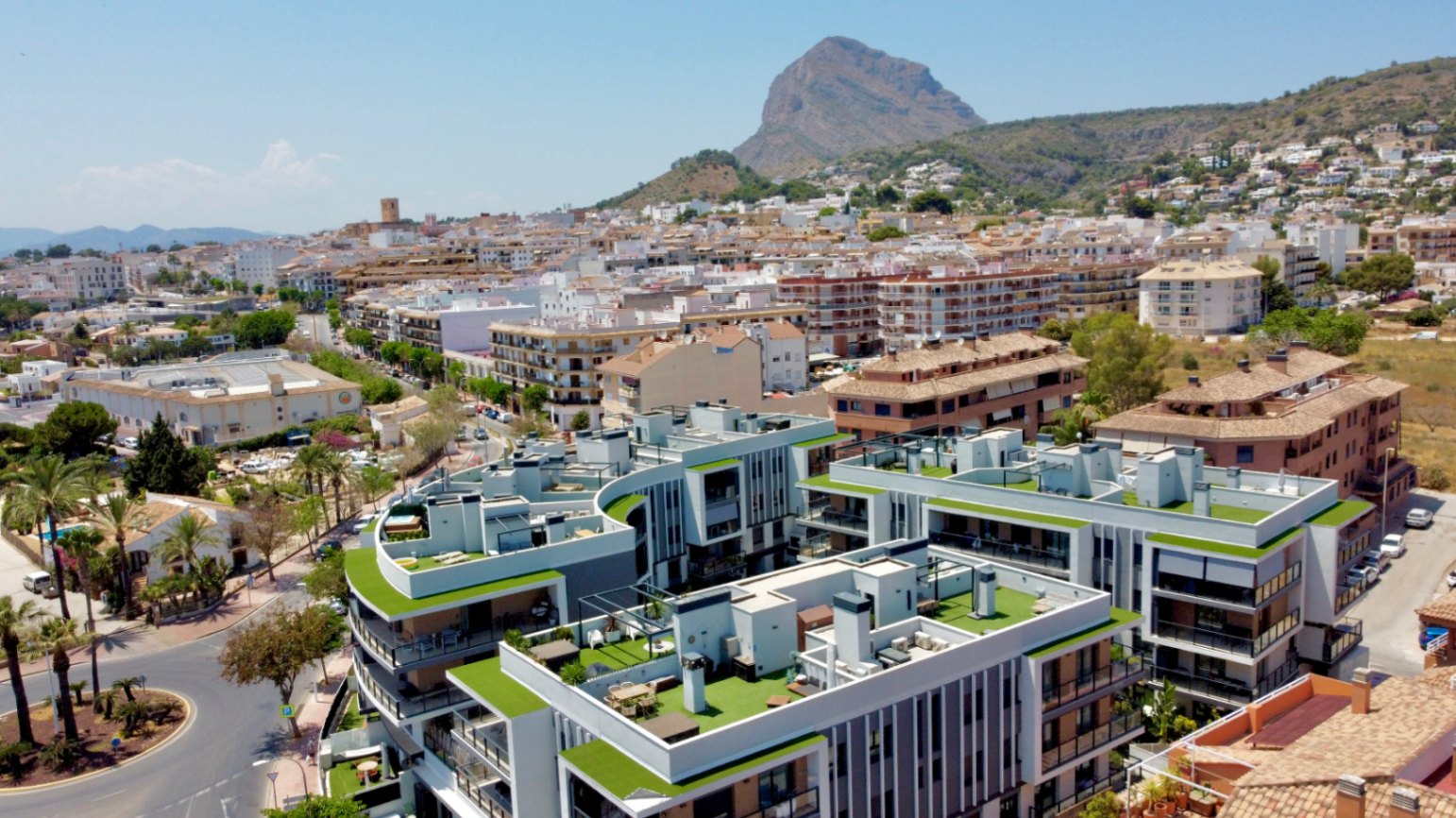 Vista aérea del complejo residencial moderno en Jávea, destacando sus azoteas ajardinadas y la cercanía al casco antiguo con el Montgó al fondo.