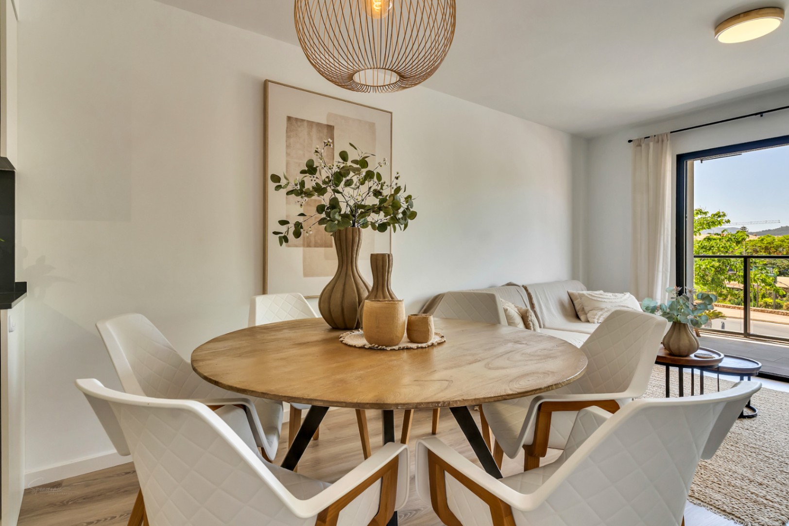 Zona de comedor con mesa circular de madera y sillas de diseño, iluminada por una lámpara de mimbre y luz natural proveniente de la terraza contigua.