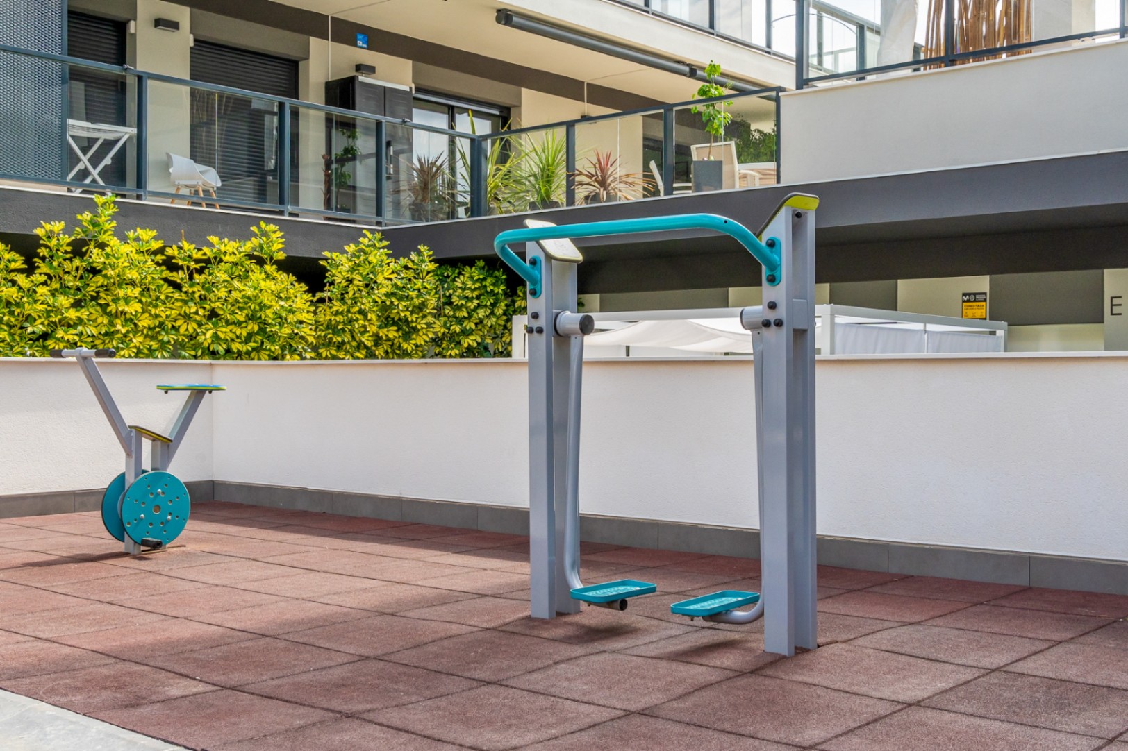 Zona de gimnasio exterior con máquinas de fitness sobre suelo técnico amortiguante, integrada en las áreas comunes con vegetación y vistas al edificio moderno.