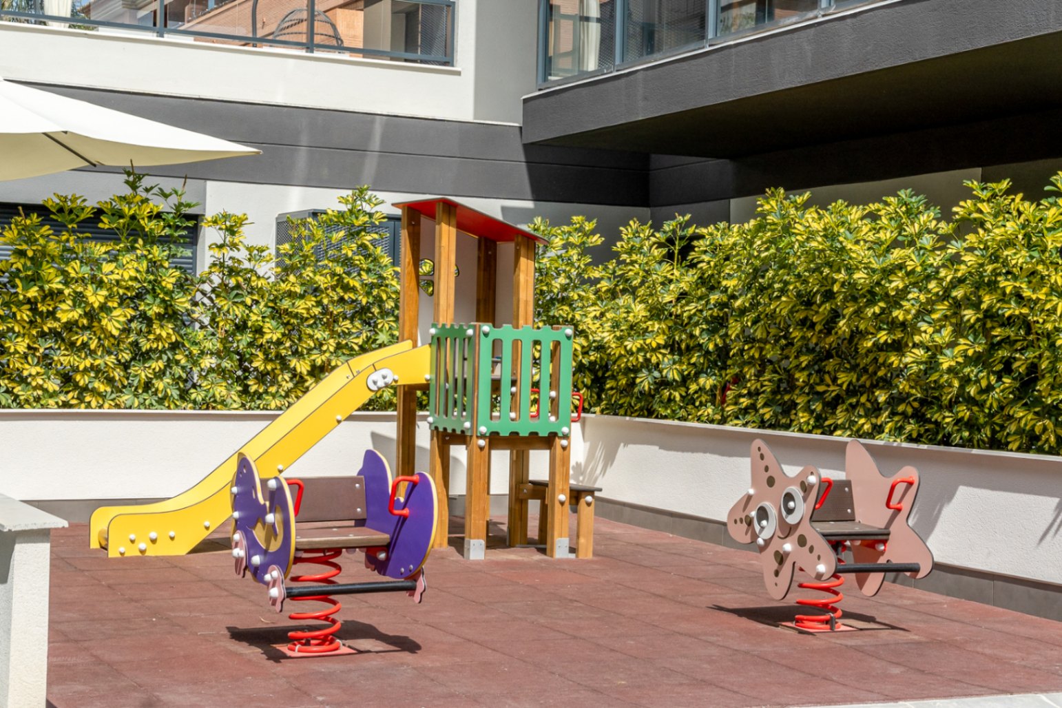 Zona de juegos infantiles comunitaria con suelo de seguridad, tobogán amarillo y balancines de muelle, rodeada de setos cuidados y arquitectura moderna.