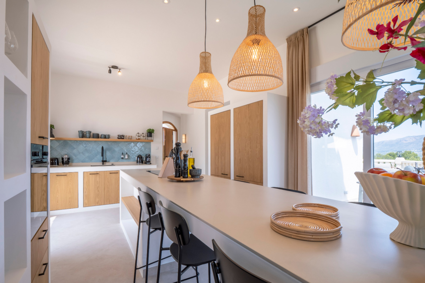 Cocina luminosa con isla central, lámparas de mimbre y mobiliario de madera clara. Azulejos turquesa en la pared.