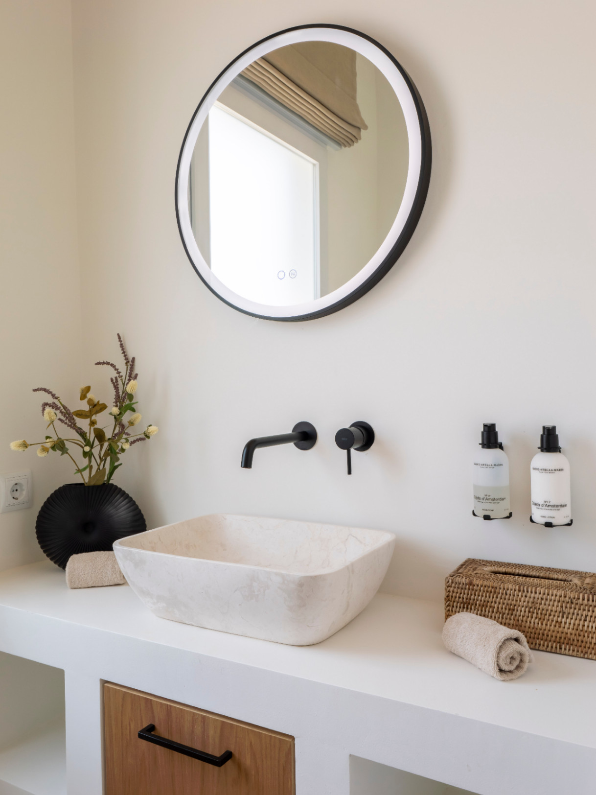 Detalle de baño con lavabo de piedra, grifería negra mate y espejo redondo con luz integrada.