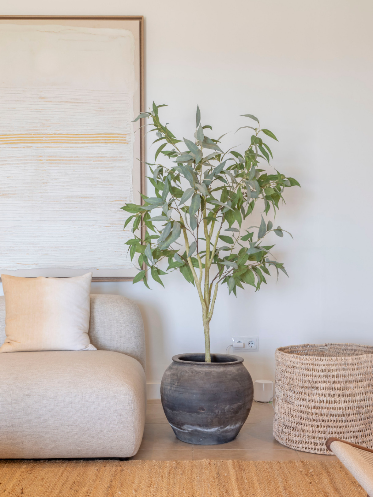 Detalle de salón con sofá beige, planta en maceta de cerámica y cesto de mimbre sobre alfombra de yute.