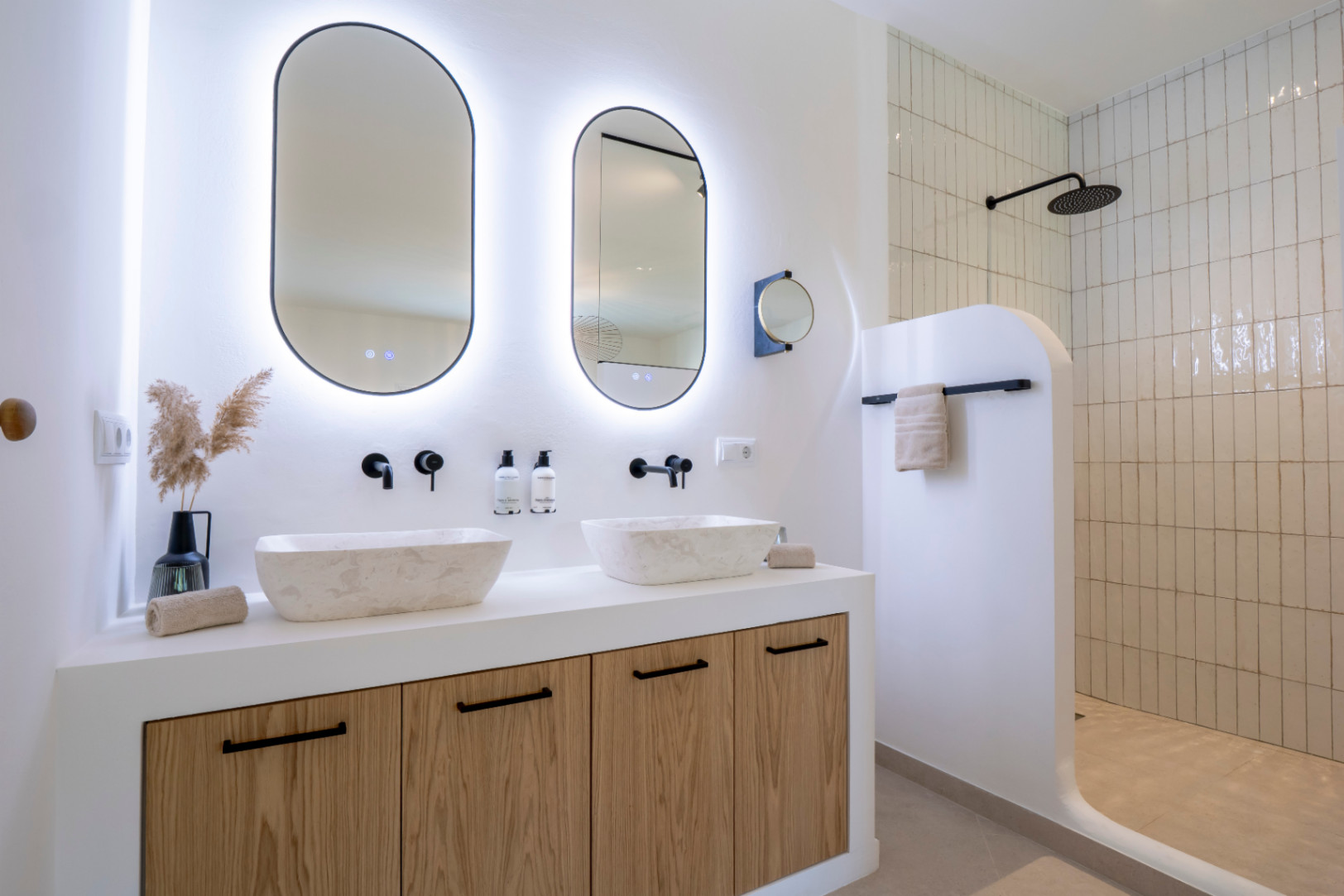 Elegante baño con lavabos de piedra, espejos ovalados con luz y ducha con azulejos verticales.