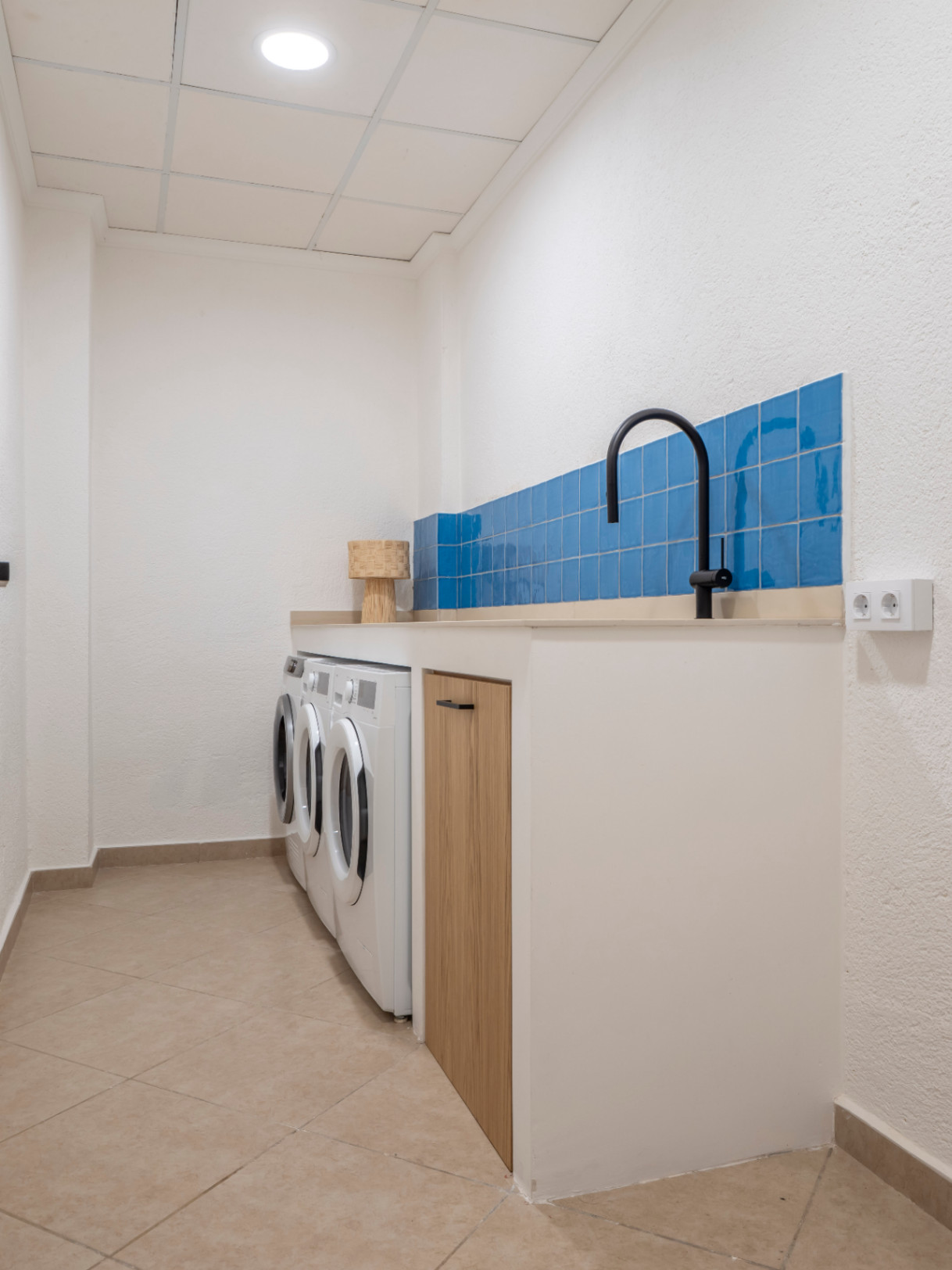 Lavandería funcional con azulejos azules, encimera beige, lavadoras blancas y suelo de baldosas.
