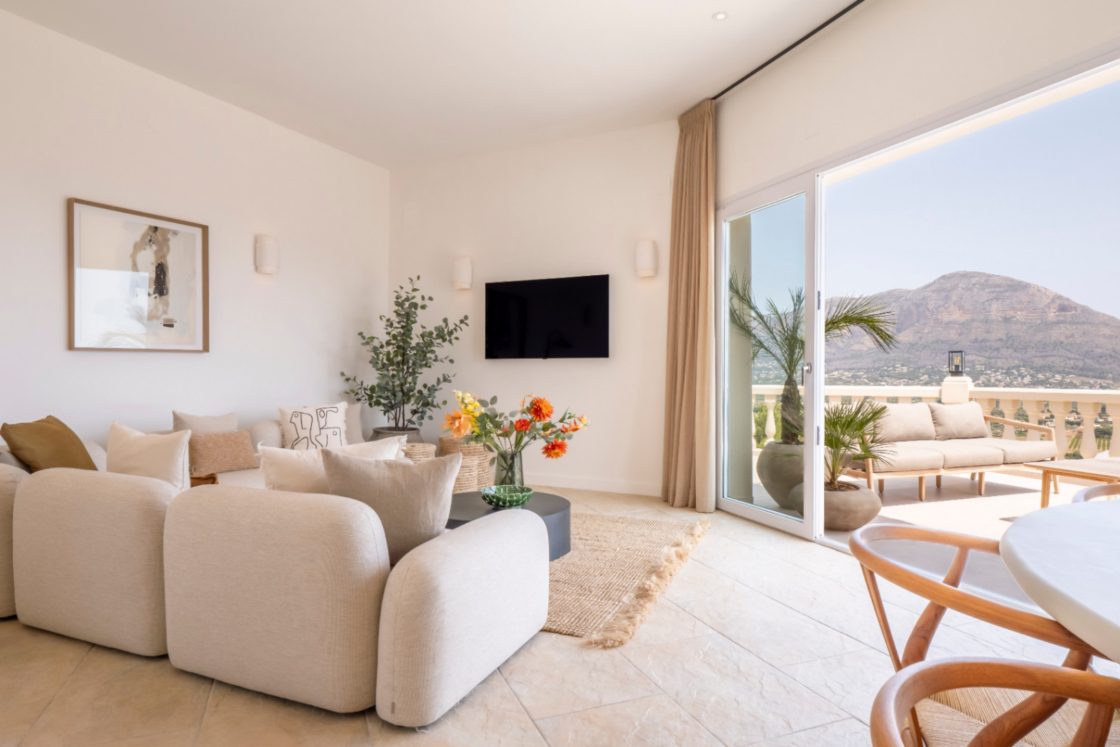Luminoso salón con acceso a terraza. Sofá modular beige, TV y decoración floral. Vistas a la montaña desde Jávea.