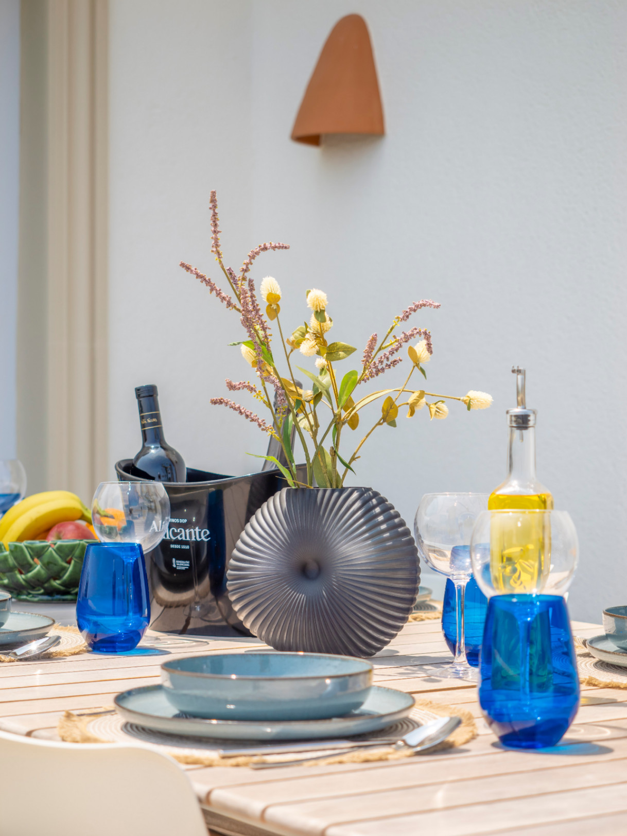 Mesa de comedor al aire libre con vajilla azul, florero gris y enfriador de vino Alicante.