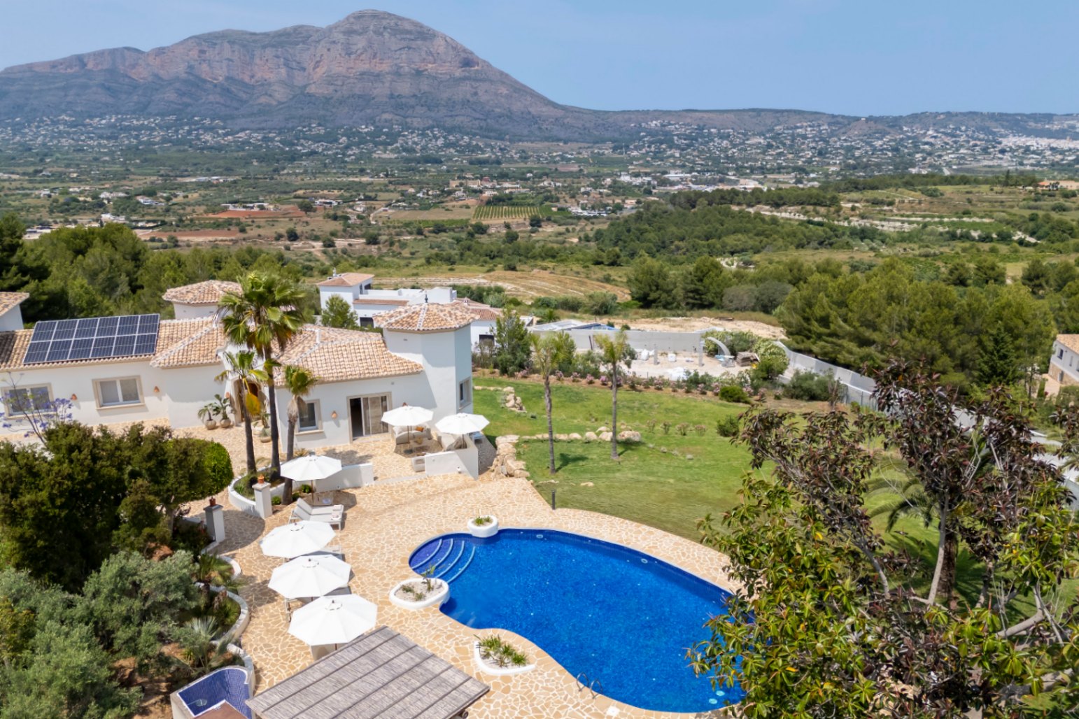 Vista aérea de la villa con piscina azul, terrazas con sombrillas blancas y jardín mediterráneo con vistas a la montaña.