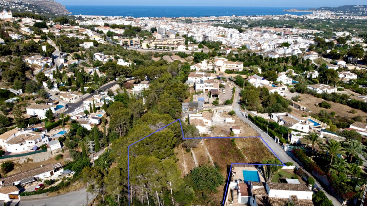 Vista aérea de la parcela delimitada en azul, destacando su vegetación autóctona y su ubicación privilegiada con vistas panorámicas al mar y al casco urbano de Jávea.