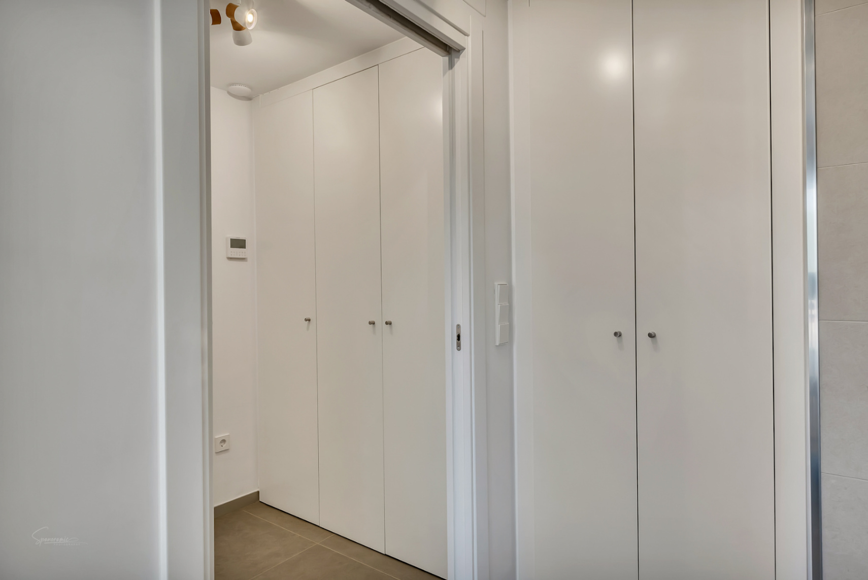 Armarios empotrados de suelo a techo con acabado blanco minimalista y tiradores discretos, integrados en un pasillo con suelos porcelánicos y panel de control digital.