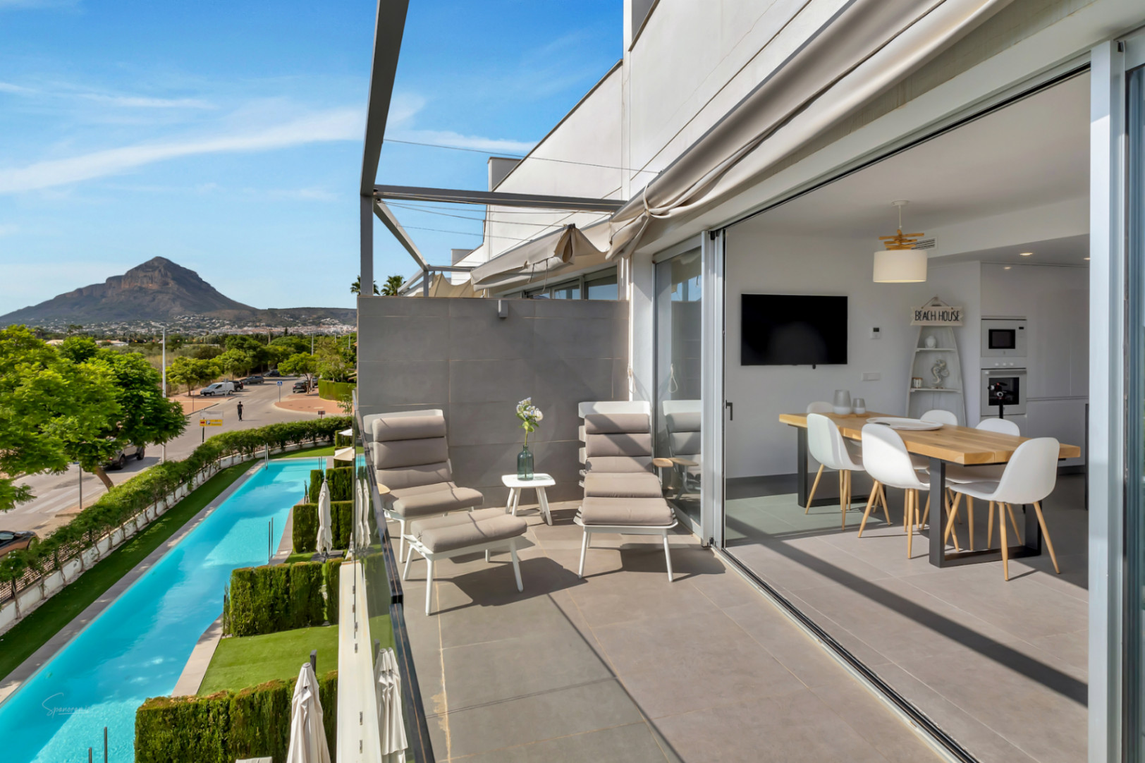Balcón con mobiliario de descanso y vistas a la piscina comunitaria y la montaña. El salón comedor se integra mediante grandes ventanales correderos.