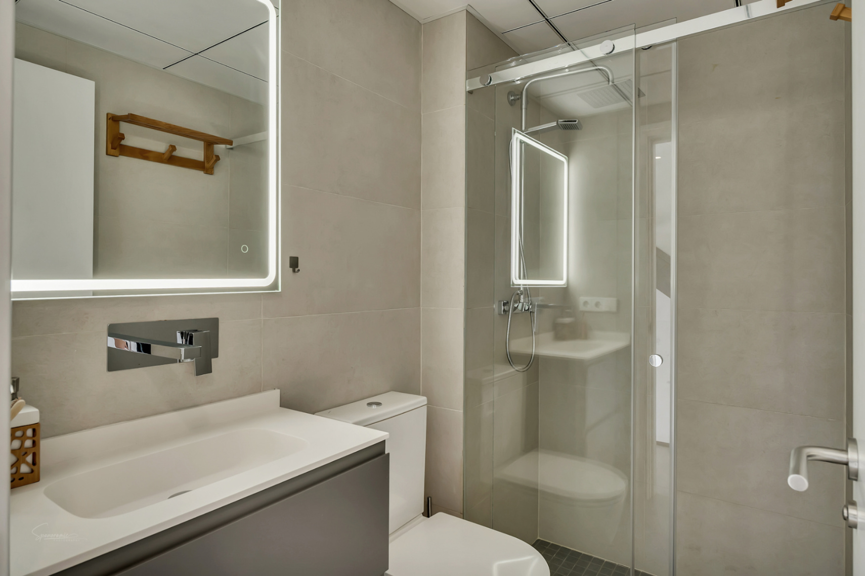 Baño moderno con azulejos en tonos neutros, espejo con iluminación LED perimetral y ducha de obra con mampara de cristal corredera.
