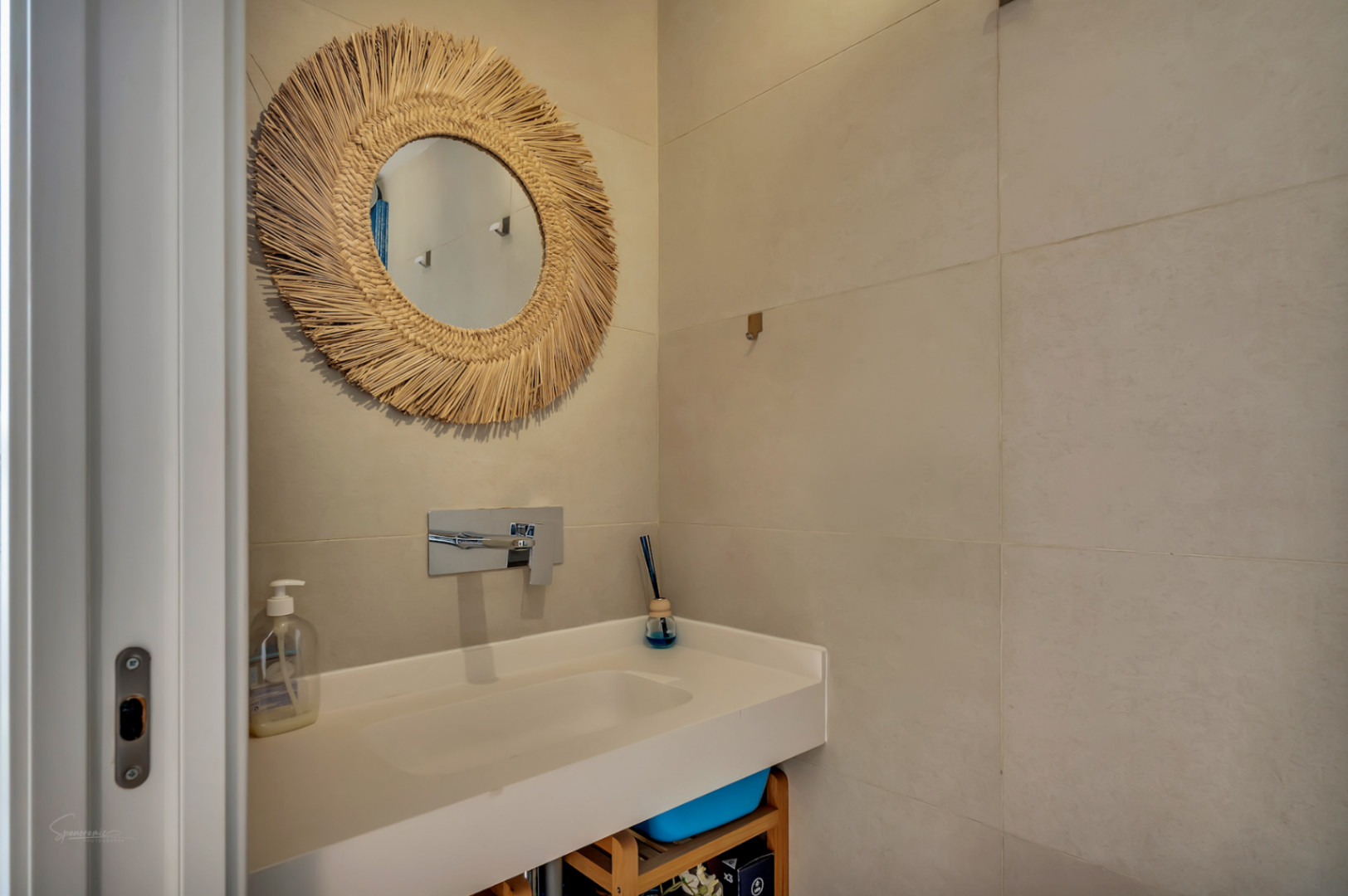 Baño moderno con lavabo integrado blanco, grifería empotrada cromada y un espejo decorativo con marco de fibras naturales sobre azulejos de gran formato en tonos arena.