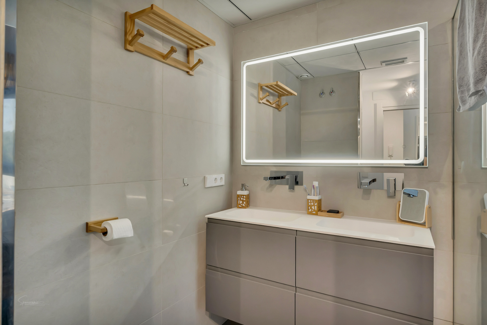 Baño moderno con mueble suspendido gris, lavabo doble integrado y un gran espejo con iluminación LED perimetral. Incluye grifería empotrada y accesorios decorativos en madera natural.
