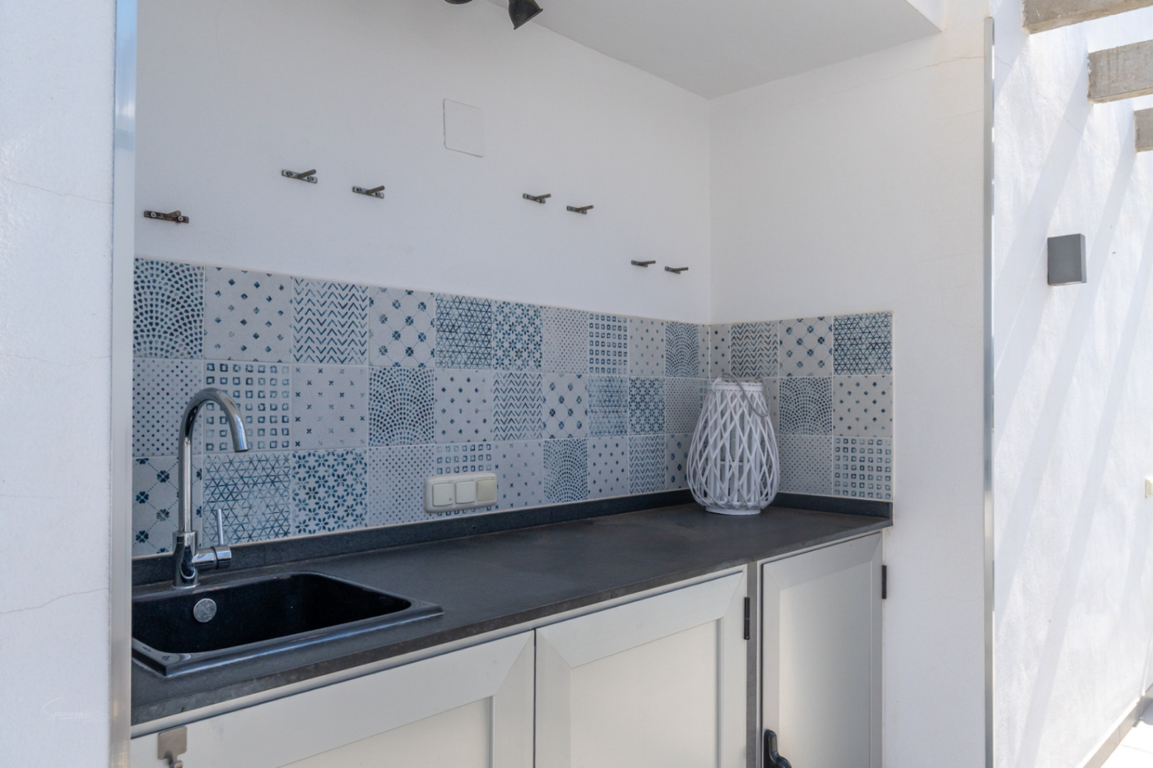 Cocina de verano exterior con encimera oscura, fregadero integrado y un elegante frontal de azulejos con motivos geométricos en tonos azules.