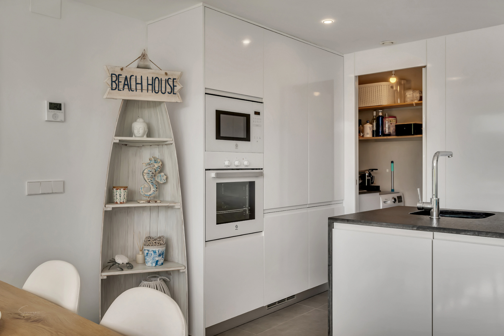 Cocina moderna con armarios blancos de alto brillo, electrodomésticos integrados y zona de lavadero adyacente con estantería decorativa náutica.