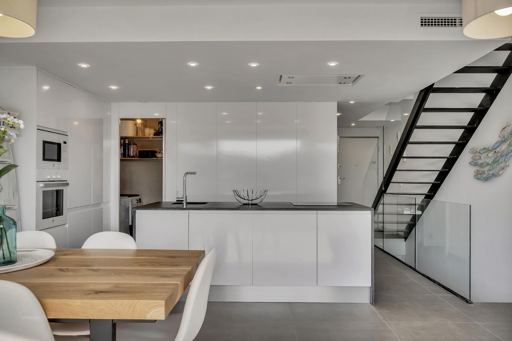 Cocina moderna de diseño minimalista con isla central, armarios blancos brillantes y lavadero integrado. La escalera de metal negro y cristal conecta con el comedor de madera.