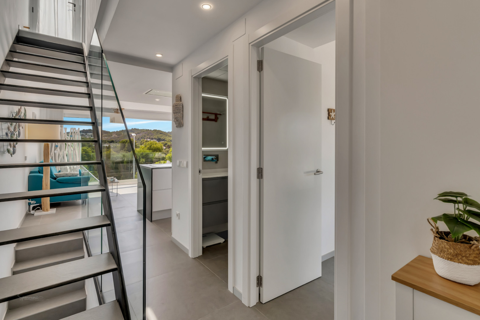 Distribuidor moderno con escalera de peldaños volados y barandilla de cristal, conectando con el salón y ofreciendo vistas despejadas al entorno natural.