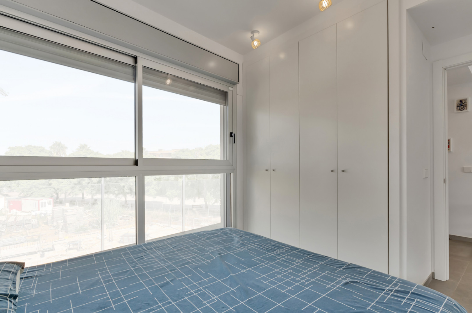 Dormitorio luminoso con armarios empotrados blancos, grandes ventanales con vistas despejadas y una iluminación moderna mediante focos orientables.