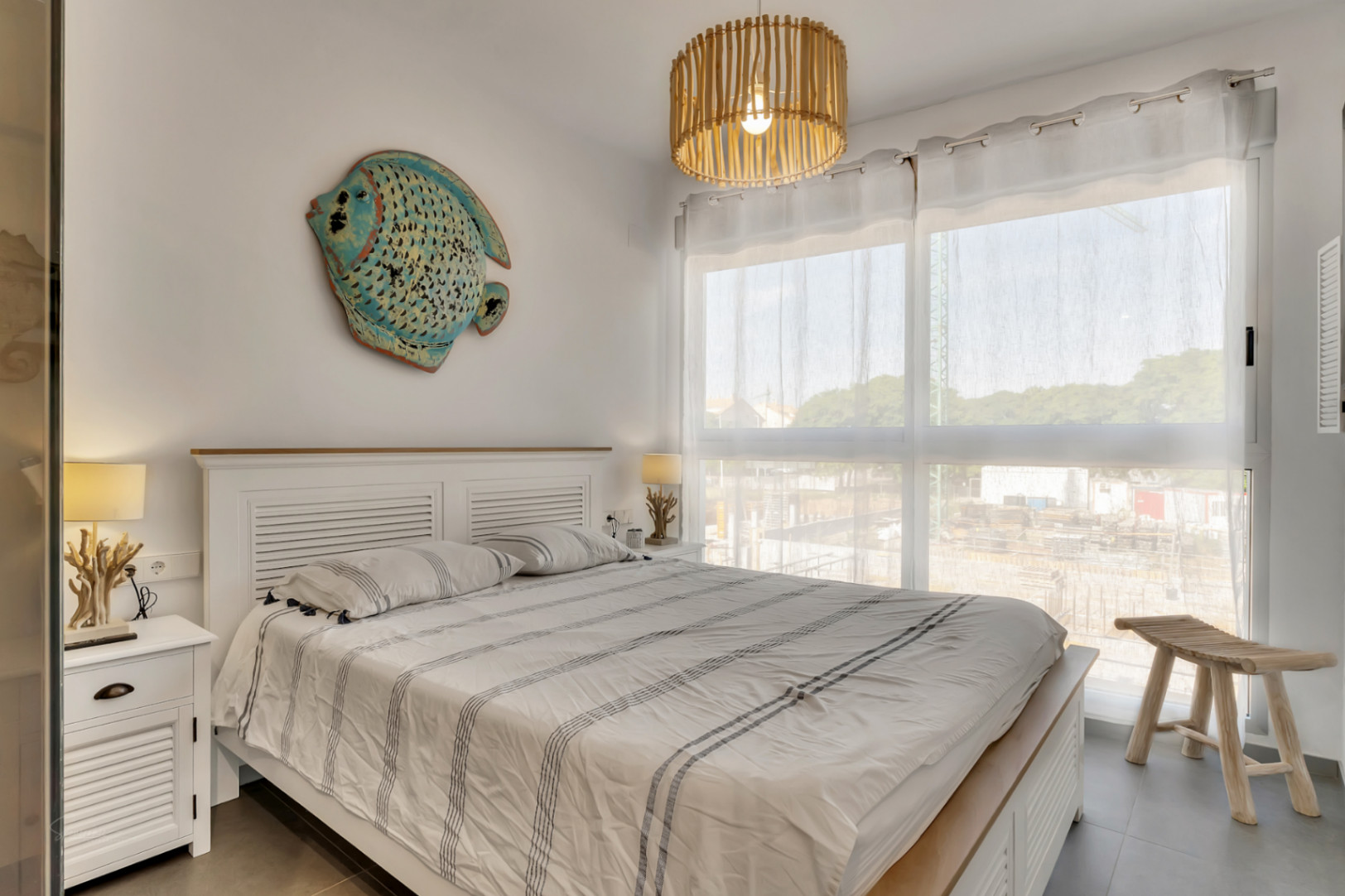 Dormitorio luminoso con cama de madera blanca, decoración de inspiración marina, lámpara de techo de bambú y gran ventanal con luz natural.