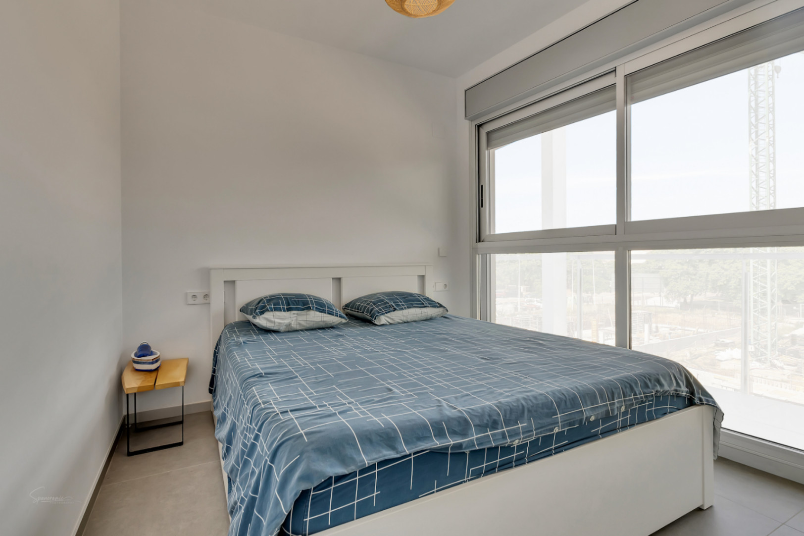 Dormitorio luminoso con cama doble, amplio ventanal de aluminio gris con persianas motorizadas y suelos de gres en tonos neutros.