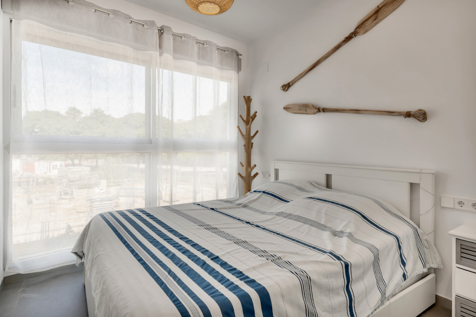 Dormitorio luminoso con decoración náutica, cama doble, ventanal con cortinas traslúcidas y detalles en madera como remos decorativos y perchero artesanal.