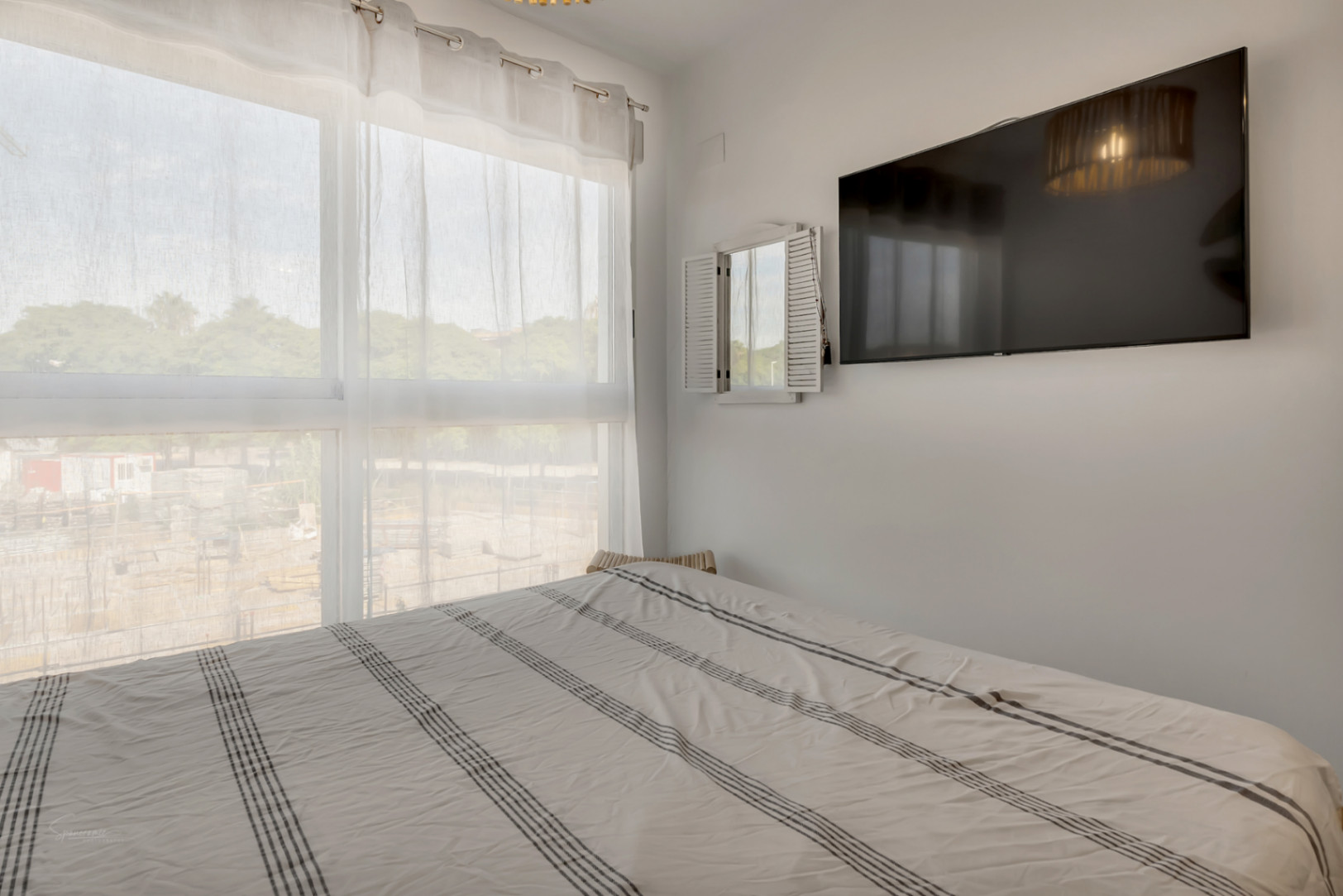 Dormitorio luminoso con gran ventanal, cortinas traslúcidas, cama doble con ropa de rayas y televisión de pantalla plana integrada en la pared blanca.
