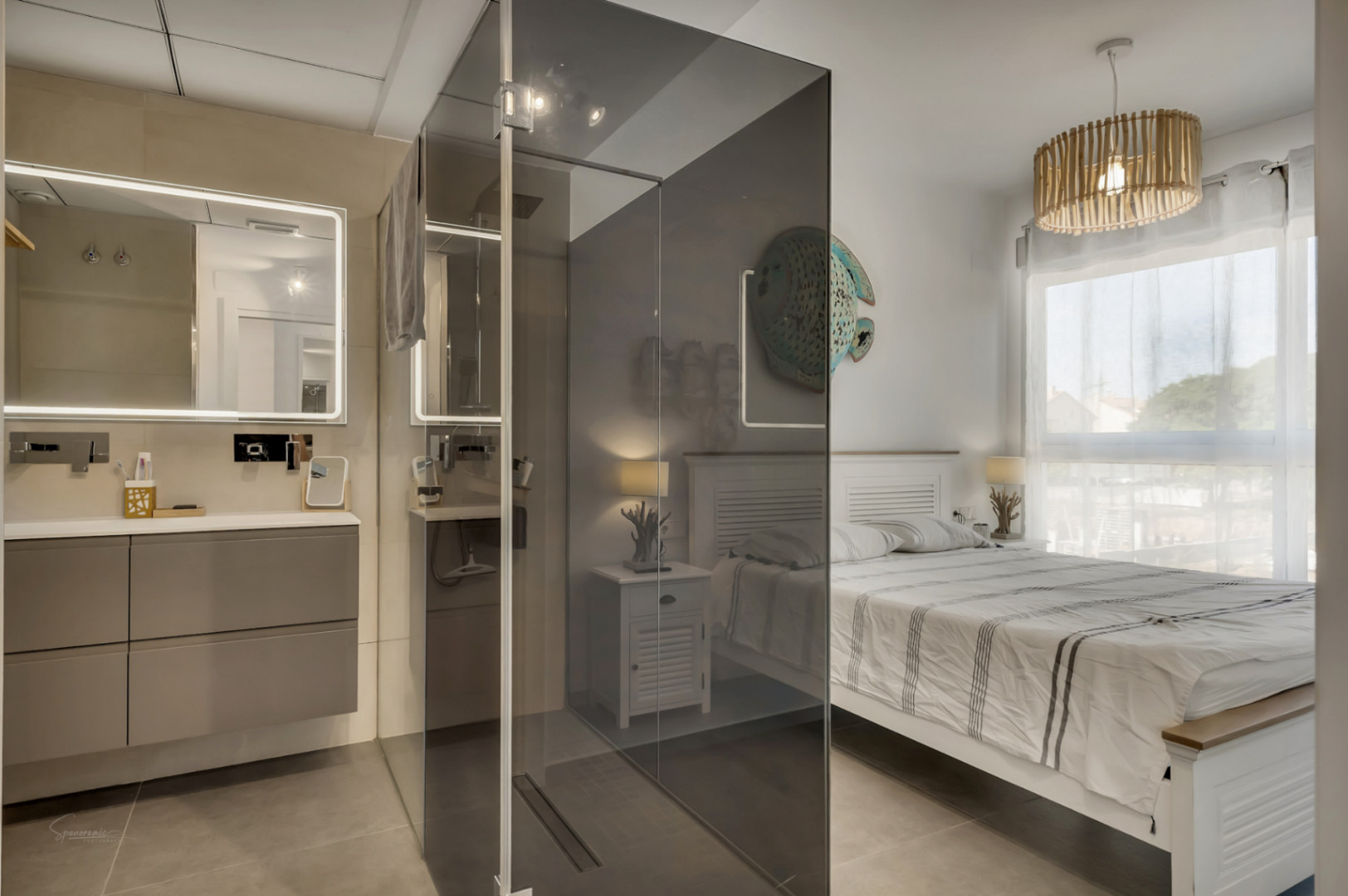 Dormitorio principal con baño integrado, separado por una mampara de cristal ahumado, lavabo suspendido con espejo retroiluminado y cama de madera blanca.