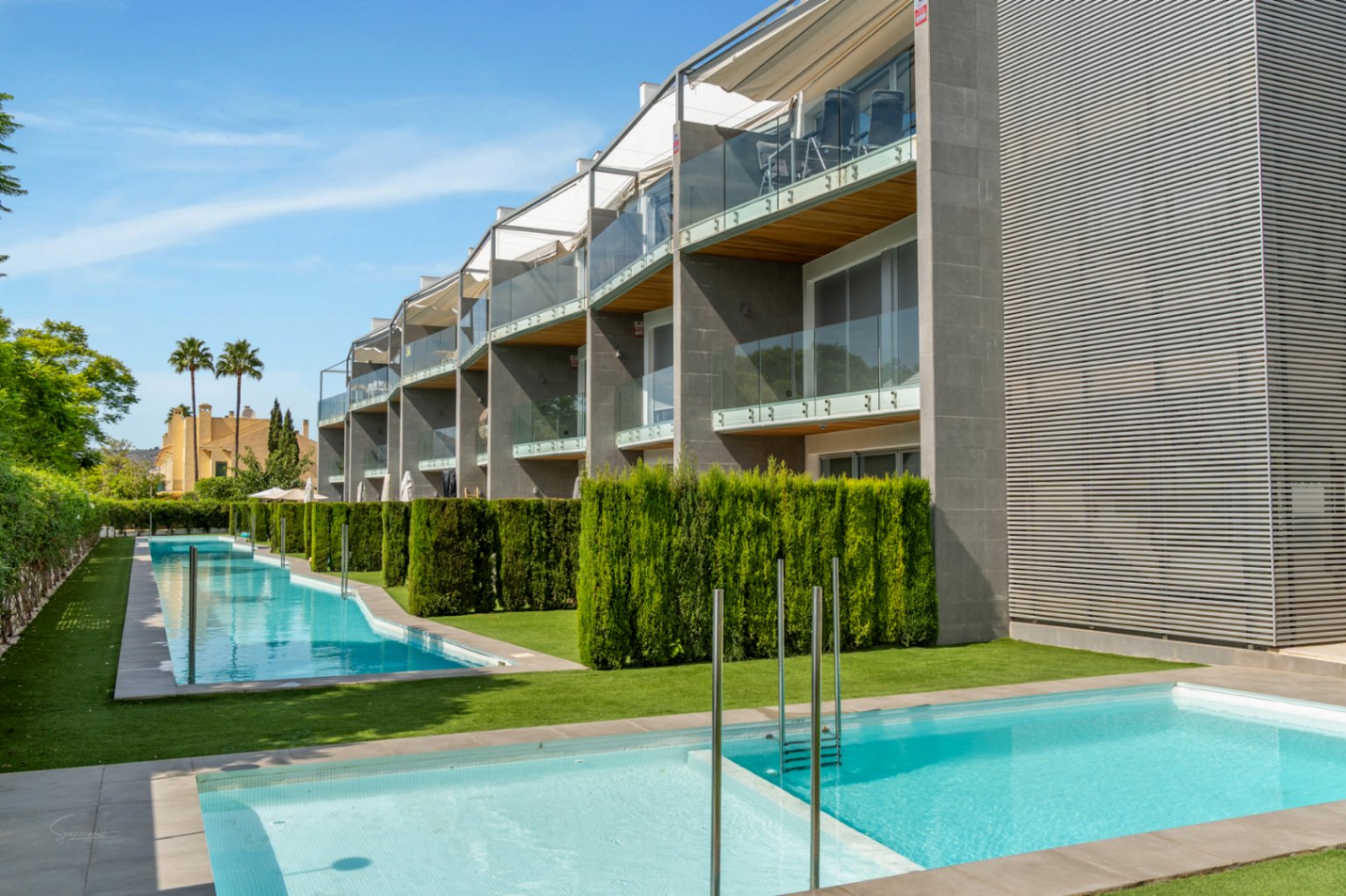 Fachada moderna con balcones de cristal y madera sobre una zona comunitaria ajardinada con dos piscinas rectangulares y setos divisorios.