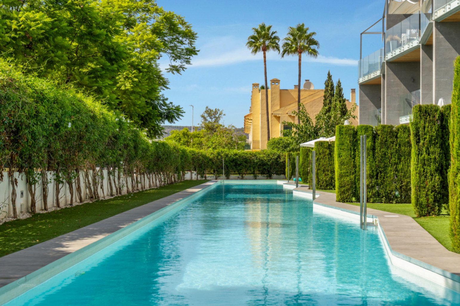 Piscina comunitaria de diseño alargado rodeada de vegetación cuidada, setos altos y zonas de césped, situada en una urbanización moderna.