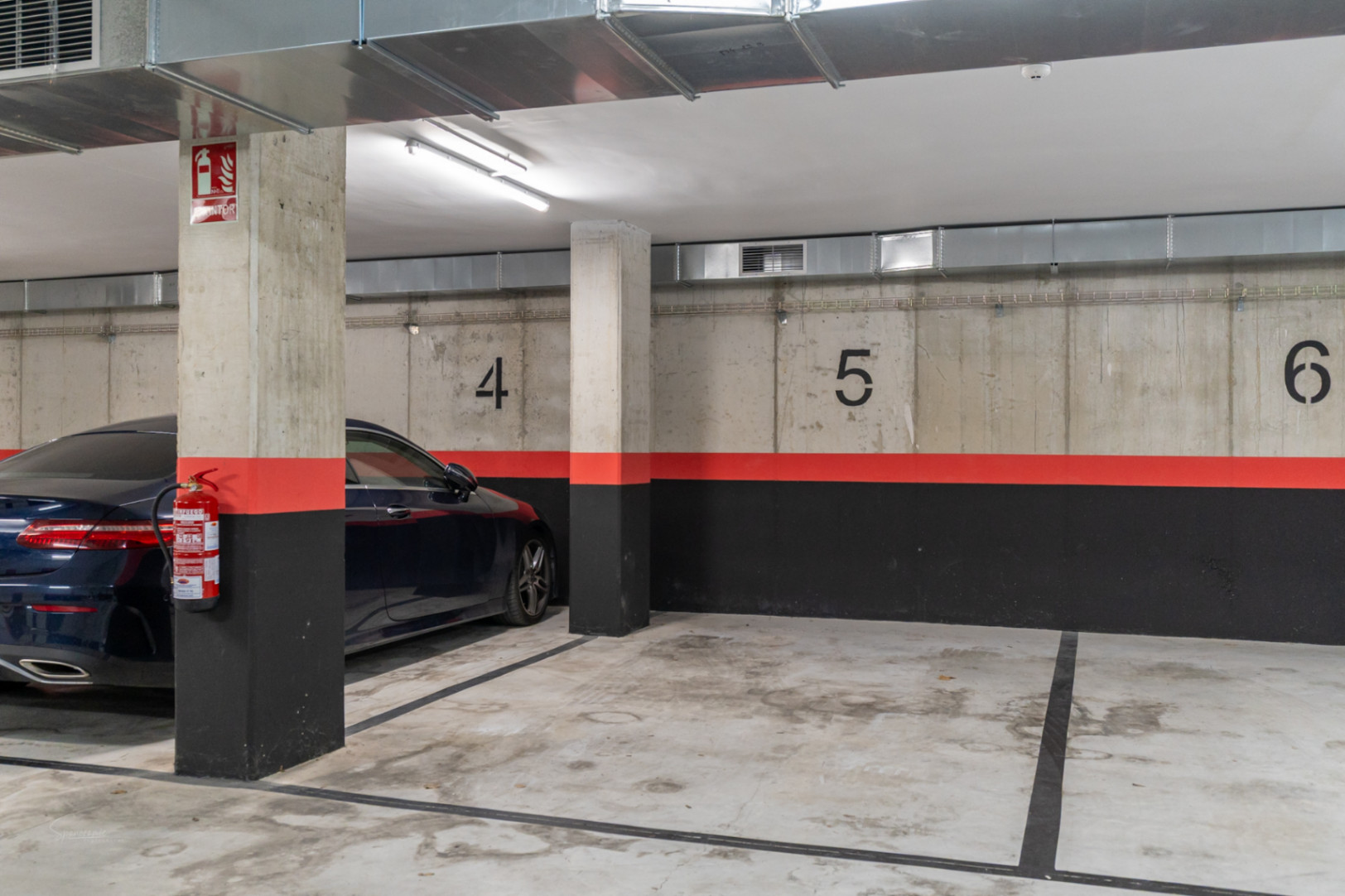 Plaza de garaje subterránea numerada en edificio moderno, con paredes de hormigón visto, conductos de ventilación y sistemas de seguridad contra incendios.