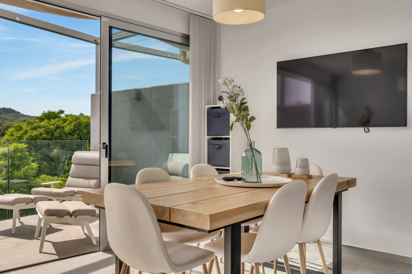 Salón comedor con mesa de madera maciza y sillas blancas, conectado mediante un gran ventanal a una terraza con mobiliario de exterior y vistas despejadas.