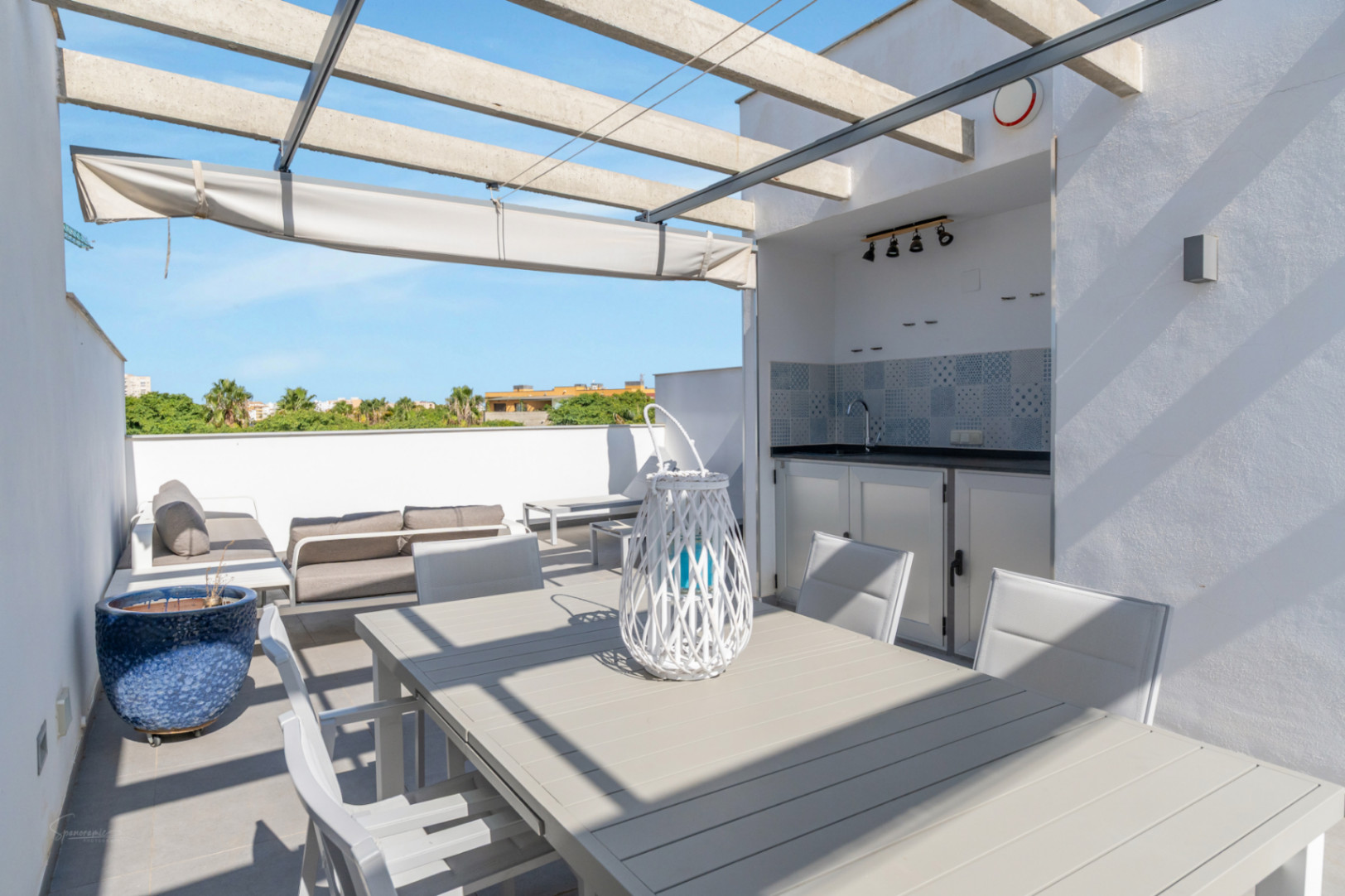 Terraza privada en la azotea con cocina de verano integrada, zona de comedor y salón exterior bajo una pérgola de hormigón con toldo retráctil.