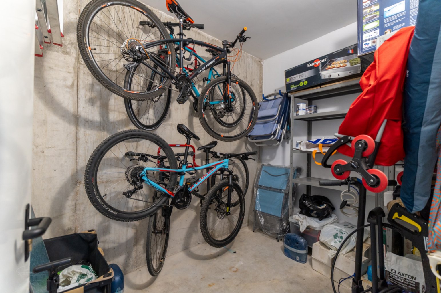 Trastero privado con paredes de hormigón visto, equipado con soportes de pared para bicicletas y estanterías metálicas para un almacenamiento eficiente.