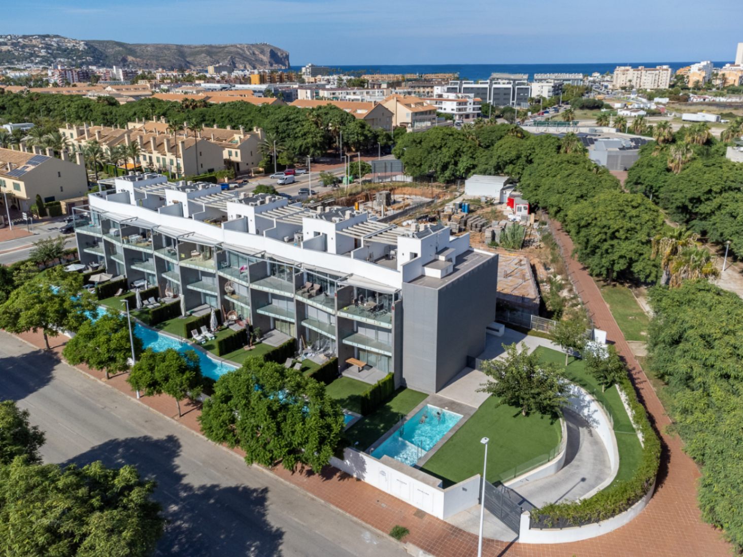 Vista aérea del complejo residencial moderno con piscinas comunitarias, jardines privados y acceso al garaje subterráneo, situado cerca de la costa con vistas al mar.