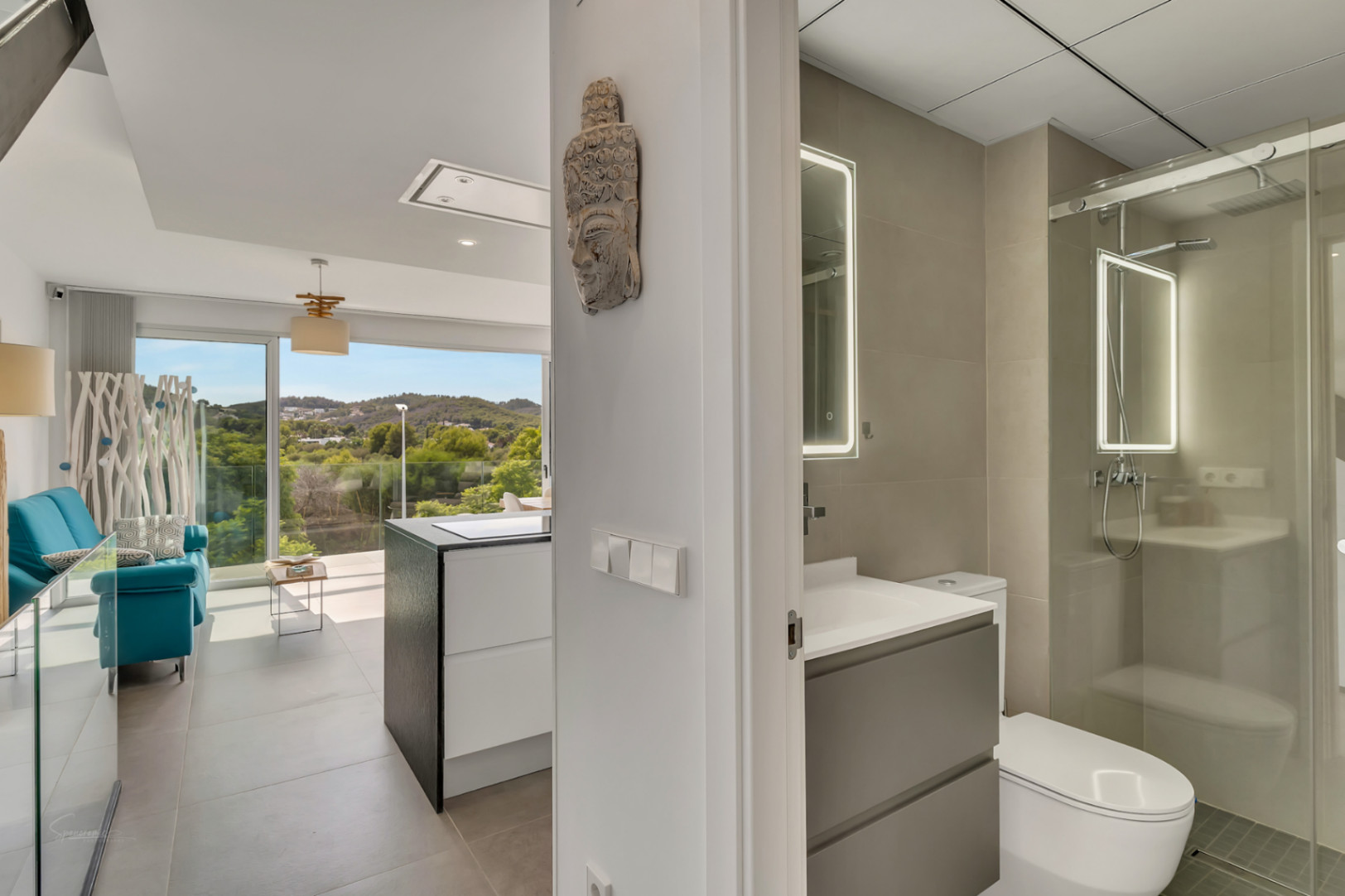 Vista desde el pasillo que muestra un baño moderno con espejo LED y ducha de cristal, junto al salón de concepto abierto con vistas panorámicas al campo.