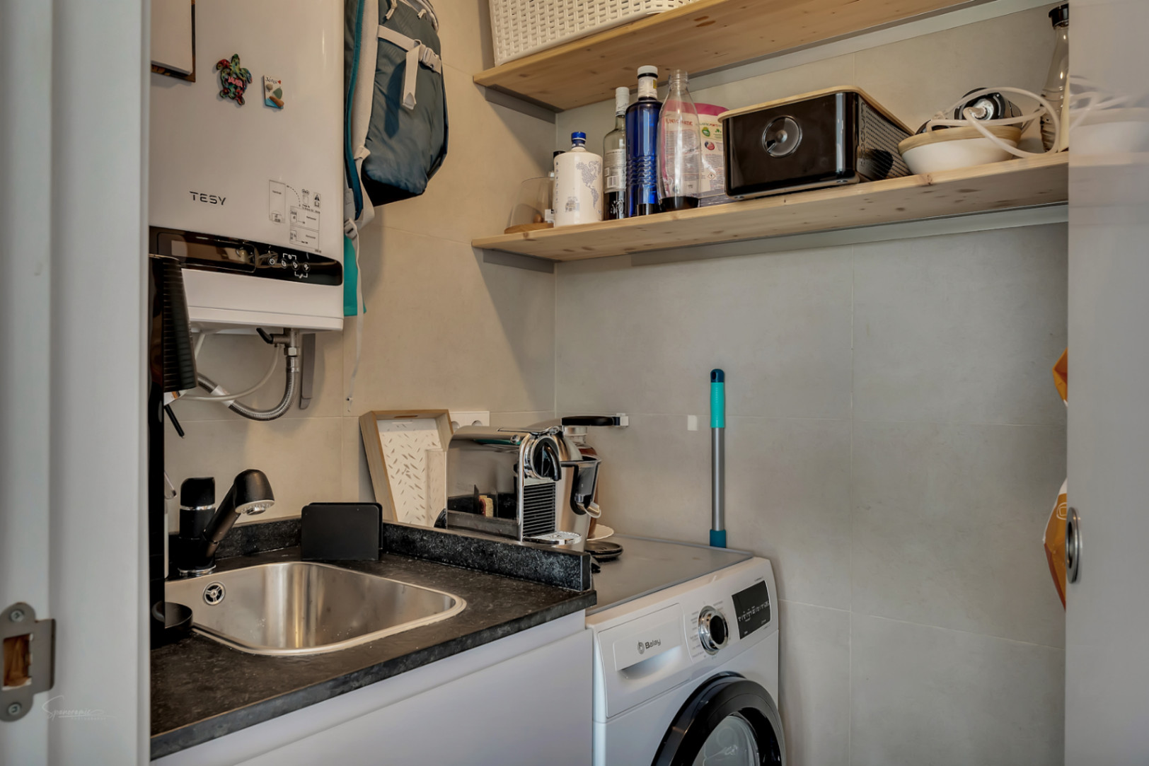 Zona de lavandería funcional con lavadora Balay, fregadero de acero inoxidable, encimera oscura y estanterías de madera natural sobre paredes de gres porcelánico.