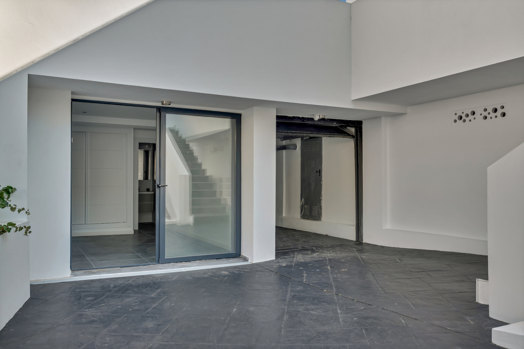 Acceso a la planta baja con entrada independiente acristalada, suelos de gres oscuro y zona de garaje o trastero integrada en la arquitectura minimalista blanca.