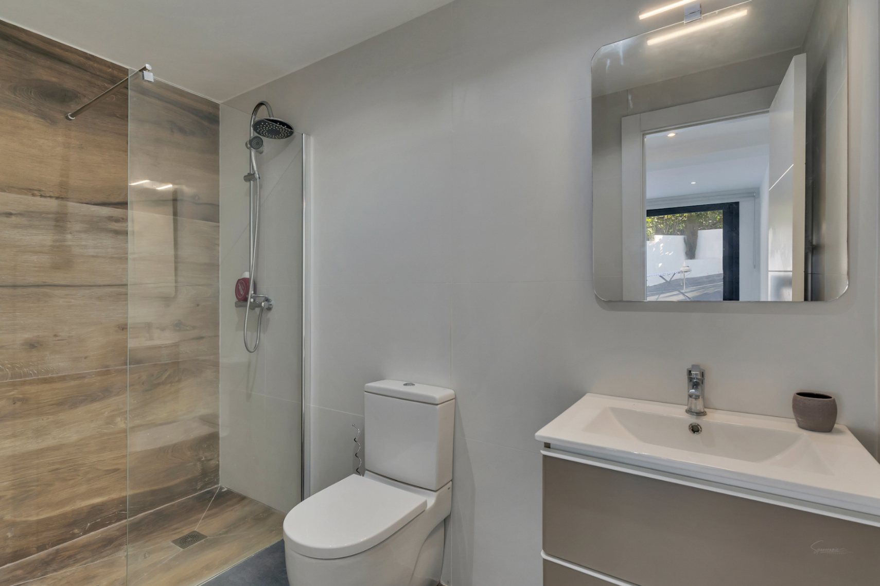 Baño moderno con ducha a ras de suelo revestida en gres efecto madera, mampara de cristal, sanitario blanco y mueble de lavabo con espejo de iluminación integrada.