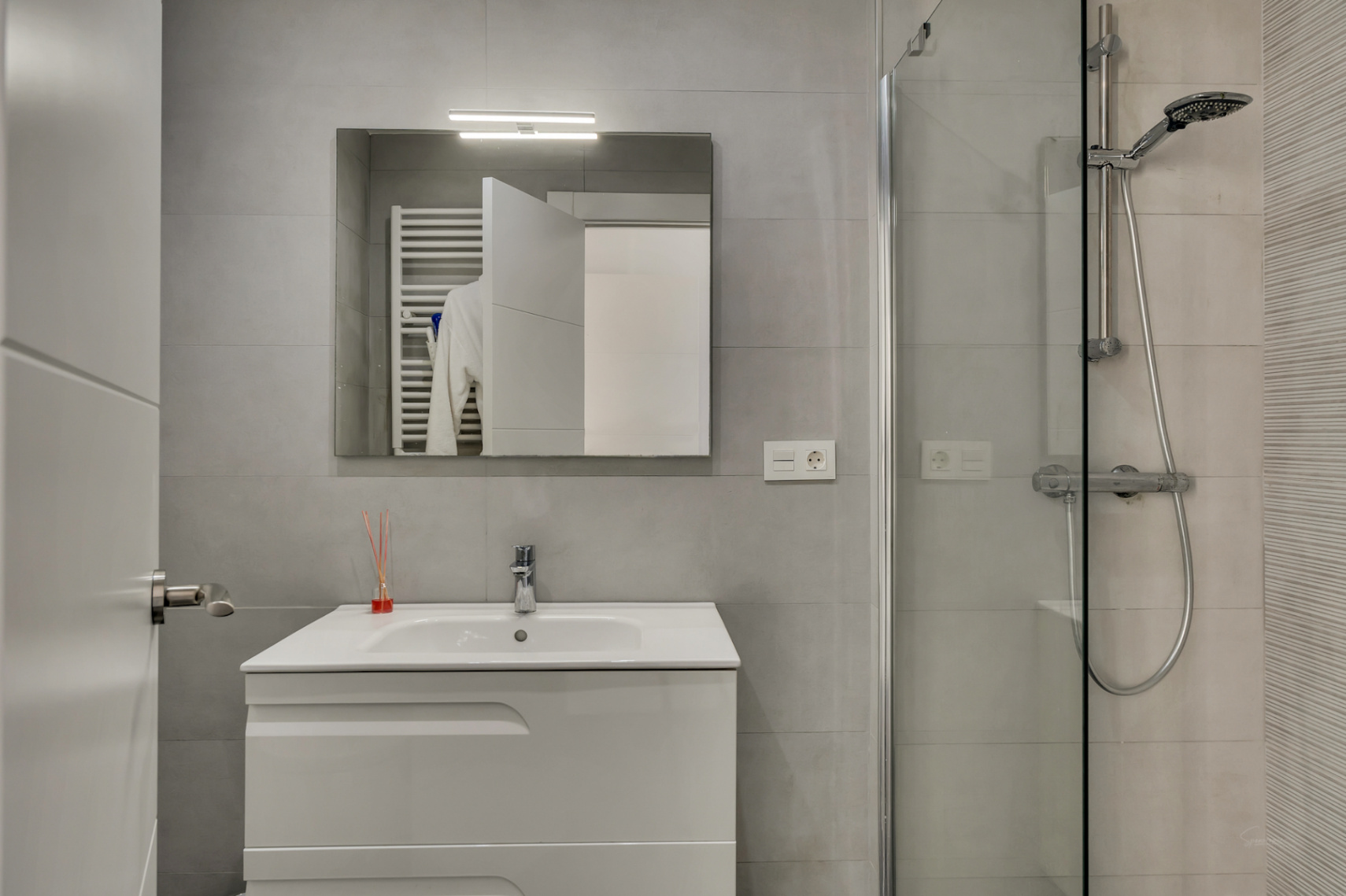 Baño moderno con revestimiento gris, mueble de lavabo blanco, espejo con iluminación superior y ducha con mampara de cristal y grifería cromada.
