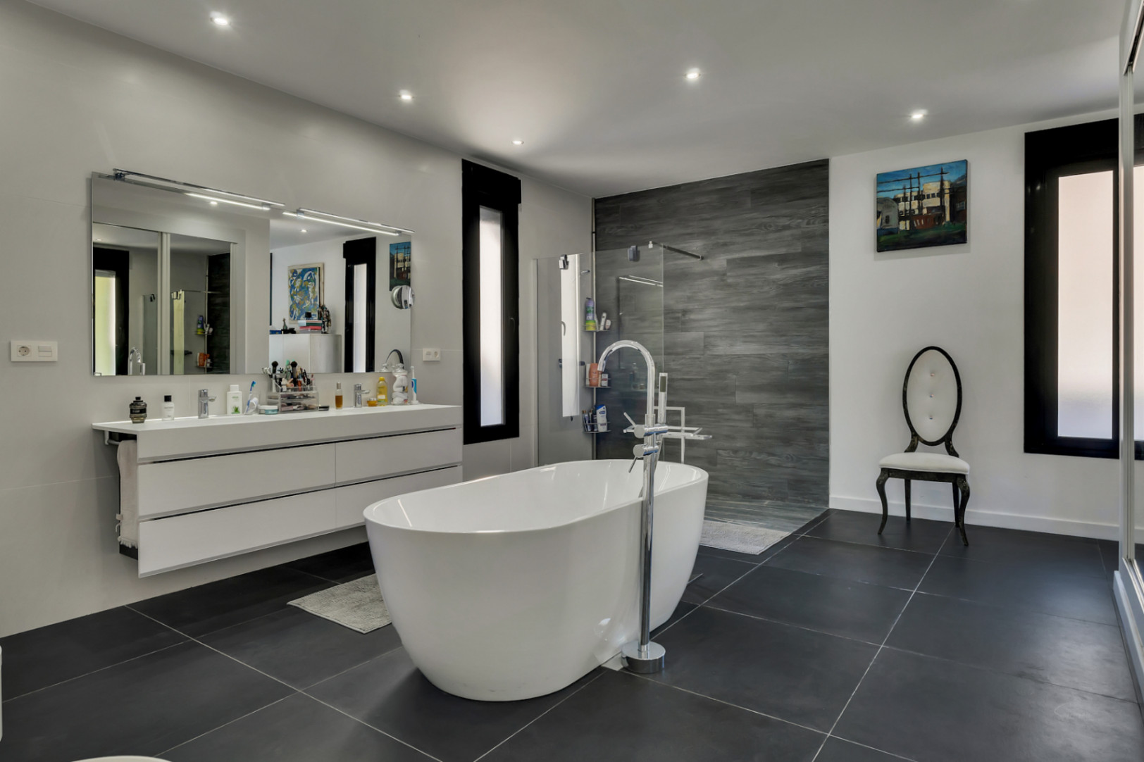 Baño principal con bañera exenta, mueble de doble lavabo blanco y ducha italiana con revestimiento de efecto madera gris sobre suelo porcelánico oscuro.
