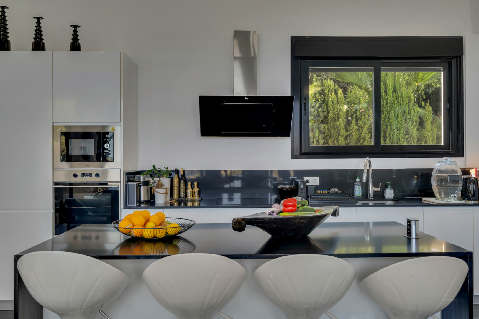 Cocina de concepto abierto con isla negra, taburetes de diseño blancos y electrodomésticos integrados frente a un ventanal con vistas al jardín.