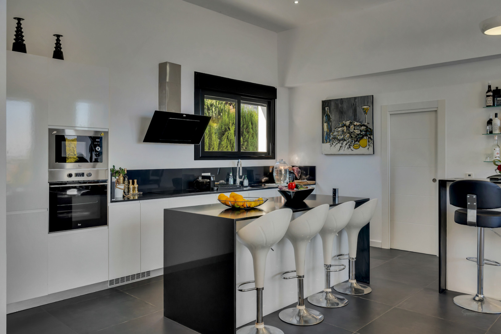 Cocina de diseño contemporáneo con isla central negra, taburetes ergonómicos blancos, electrodomésticos integrados y ventanal con marcos negros.