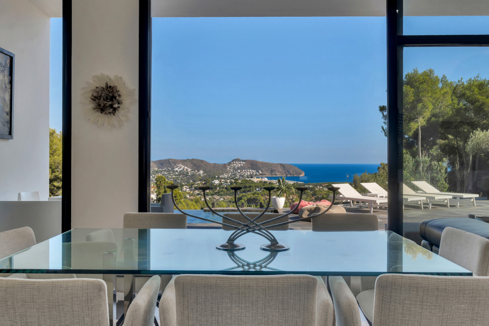 Comedor con mesa de cristal y vistas panorámicas al mar y la costa de Moraira a través de grandes ventanales de suelo a techo.